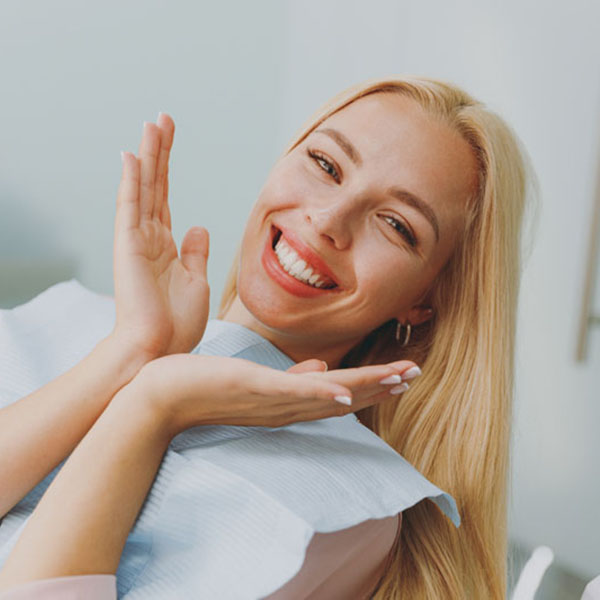 Close up young amazed smiling happy woman covered by napkin spread hands sit at dental office chair indoor light modern cabinet wait stomatologist for oral procedure Healthcare caries enamel treatment Tooth restoration